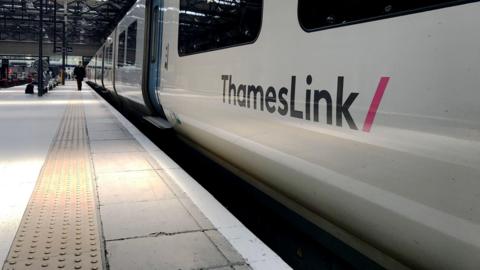 A white Thameslink train is at a platform. Several people are walking on the platform on the left.