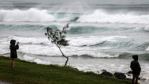 Two people on the shore watch waves crashing with sea spray in the air 