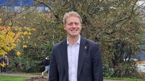 Ben Spencer, smiling and looking at the camera. Trees in the background. People walking along a path.