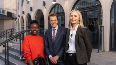 Three people stand outside the Birmingham Rep theatre: a woman in a red jumper, a man in a blue suit and glasses, and a blonde woman with a grey suit jacket