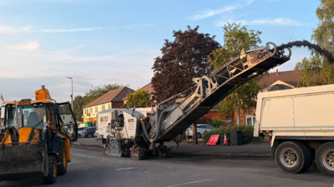 A yellow mechanical digger on a road alongside a beige truck and another machine pouring a black material into the back of it