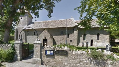 A small grey stone church with a wall in the foreground. There are trees in the church yard and the sky is blue.