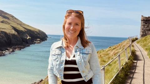 Laura, a woman with red hair, wears a denim jacket and stripy top in front of the Manx coastline on a sunny day, she has a bright smile.