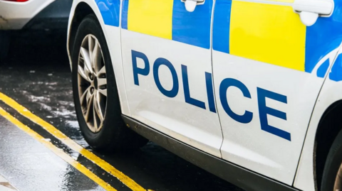 The side of a police car with "police" written down the side of it parked on double yello lines with rain falling around it 