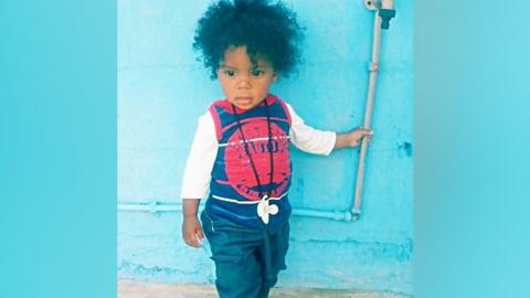 A young child in dark blue trousers and white top under a blue vest.
