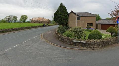 Junction of Ribchester Road and Hothersall Lane in Ribchester. It shows Hothersall Lane road sign and low stone wall in the garden of a house with a large tree to the left and people walking on the pavement of Ribchester Road with a house having a new roof being built in the background.