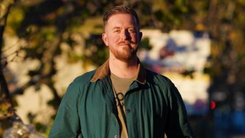 A young man with short ginger hair and a moustache and beard stands with his hands in his jacket pockets and smiling on a bridge. 