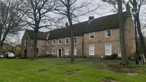 The two-storey Kepier Hall. It is a beige stone building with white windows and several trees in front of it. A car is parked on the side.