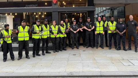 A picture of some of the security guards, there are more than 10 of them standing outside a coffee shop in Bedford, and there are three policeman with them. Most of the security guards are wearing high vis jackets.