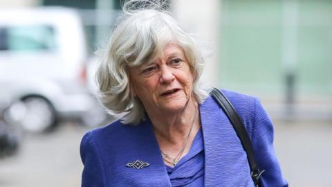 a grey-haired woman is pictured on a street. She is wearing a purple jacket and top