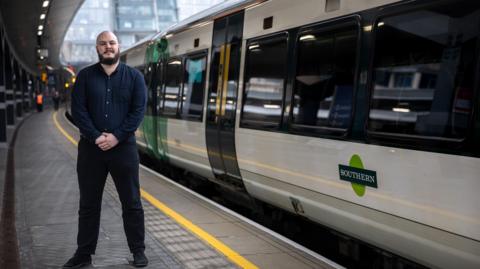 Ross Evans, wearing a blue shirt and black trousers, standing in front of a Southern Train on a platform at London Bridge station.