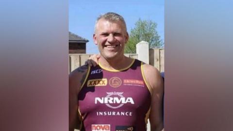 A man with short grey hair looks directly at the camera, wearing a burgundy running vest with yellow trim. The vest has sponsorship logos on the front. It is a sunny day and there is a wooden panelled fence and a tree behind him.