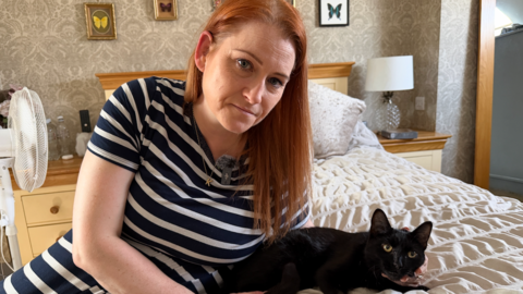 Kelly Burns is sat on a bed with a black cat. Kelly is wearing a blue and white striped dress. She has medium-length ginger hair. She is holding the black cat's face with her left hand.