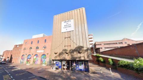 The theatre is a large orange brick building with multiple sections set back from one another and a ramp to the right side. There is a sign above the glass entrance doors which reads Prince of Wales Centre. The section to the left of the entrance has a number of archways containing colourful murals.