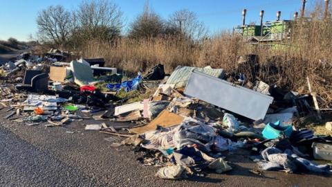 Items including sofas, mattresses and chairs littered along the lane close to Immingham tip. Trees and a hedge can be seen in the background.