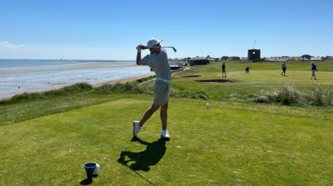 Josef has swung his golf club on a green with others playing the background and to the left there is a beach and seaside. Josef is wearing white shoes, cream shorts and t-shirt and a white cap.