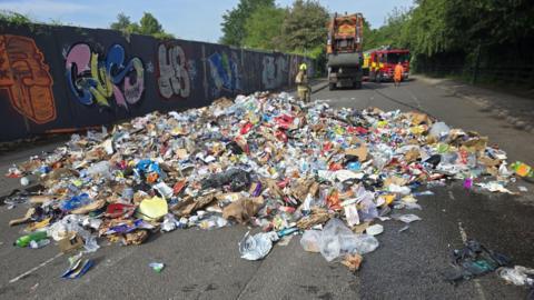 Six tonnes of rubbish has been emptied from a bin lorry all over a road after a fire took hold in the back of a bin lorry. You can see the collection vehicle in the background with a fire engine parked behind it. There is one firefighter standing behind the rubbish and a refuse collector in orange in front of the fire engine. 