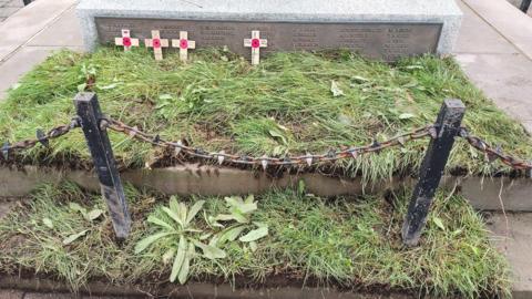 A patch of turf with overgrown grass covered in weeds laid on the bottom two steps of a war memorial. There are four wooden crosses with a poppy leaning against the plinth, where names of the fallen are engraved.