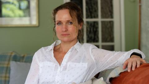 Rhianon Bragg is sat on a sofa in a white blouse. She has blue eyes and dark hair tied back, with a curly blonde strand hanging over the left hand side of her face.