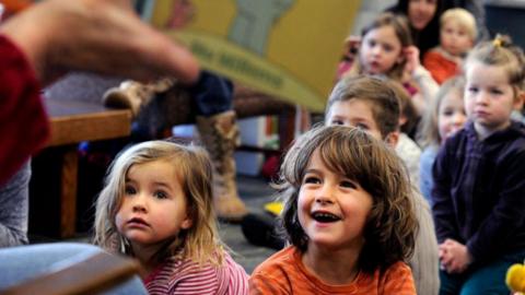 A group of children are sat on the floor engrossed by a story. The reader is holding out the open book so the kids can see a picture inside. A little girl with brown hair in the front row had a wide, open-mouthed smile.