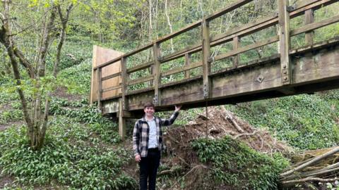 A picture of Josh Rutty stood below the bridge. He is wearing black trousers, a white top and a chequered jacket. He is stood in the middle of the woodland.