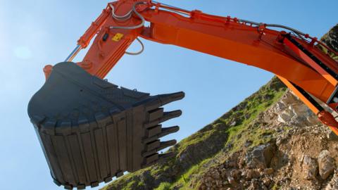 Close-up shot of a large orange excavator arm with a heavy-duty bucket. The excavator is on a hillside with blue sky in the background.
