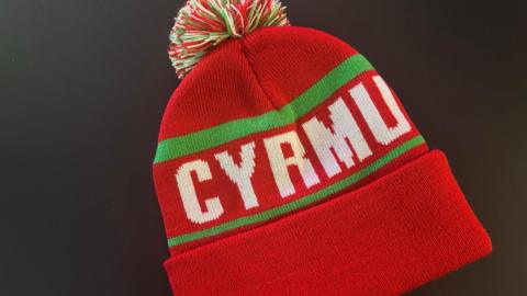 A red bobble hat which has Cyrmu in white writing across the middle, which is a misspelling of the word Cymru.