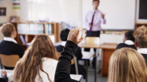 Pupils with their back to the camera sit in a classroom. The two nearest have long hair past their shoulders and the nearest one has their hand in the air. A man teaches at the front of the class as they sit behind desks.