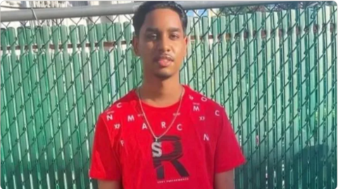 A young man wearing a red T-shirt and a silver chain around his neck, standing against a green fence