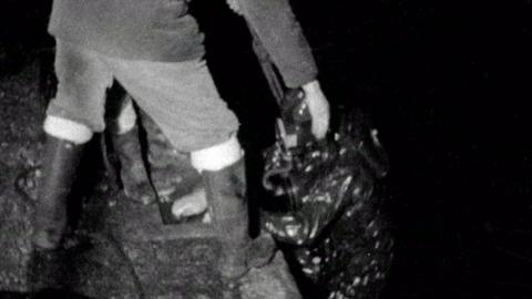 Black and white image of diver (in heavy deep diving gear), climbing back up on to dock.  Another man in waterproof boots is reaching down to help lift him back up.