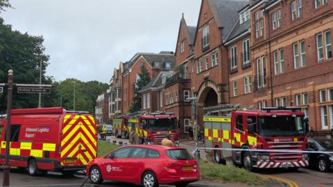Multiple fire engines and red fire service support vehicles pictured at a roadside. There is a large brick building nearby and a line of police tape.