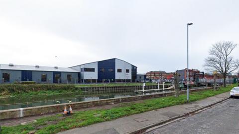 A view of the River Witham at Stamp End with a road and grass verge in the foreground and warehouses on the far bank