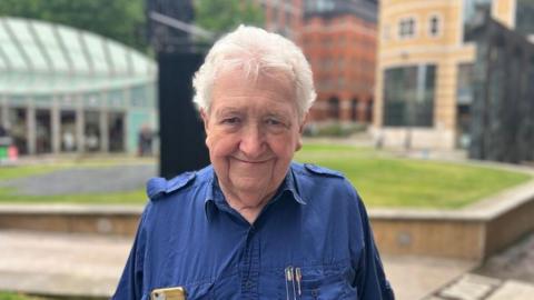 A man with white hair is smiling at the camera. He is wearing a navy blue shirt with his gold mobile phone in one breast pocket and some pens in another.