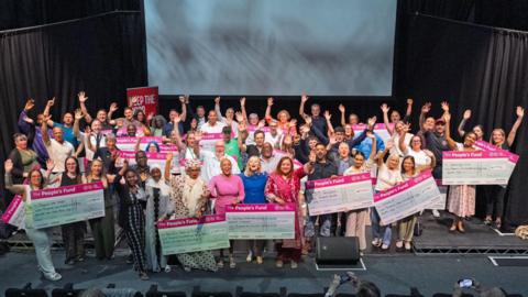 A large group of people on a stage holding giant pink cheques and raising their hands in the air