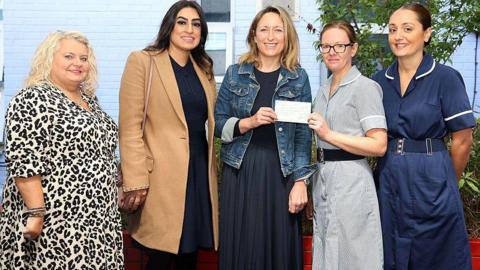 Five women stand in a line in a garden. The two on the far right are wearing nursing uniforms while the other three are dressed more casually. The woman in the centre, wearing a blue top, a blue skirt, and a blue denim jacket, is handing a cheque to one of the nurses while smiling at the camera.