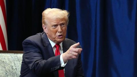 An image of US President Donald Trump, who is sat on a chair and wearing a black suit jacket, white shirt and red tie. He is pointing. 
