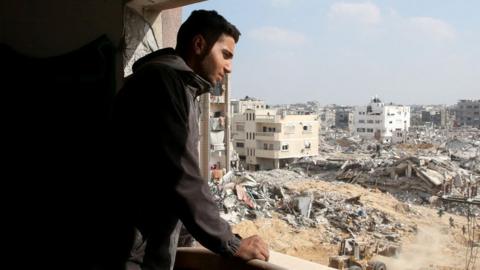 A man looks across a landscape of damaged buildings from a window in Gaza