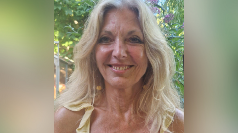 A smiling Lynn Knapp outside by a tree. Her blonde hair is down.
