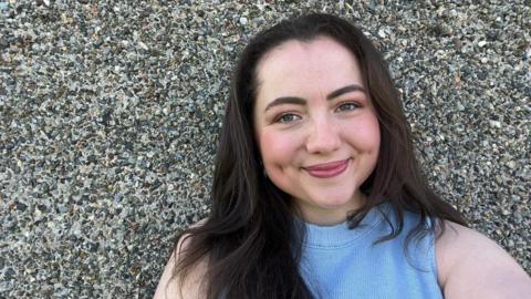 Llun o Erin Hughes yn sefyll y tu allan i wal ei thy. Mae ganddi wallt hir tywyll ac mae hi'n gwenu ar y camera ar ffurf hunlun. Mae hi'n gwisgo top lliw glas.