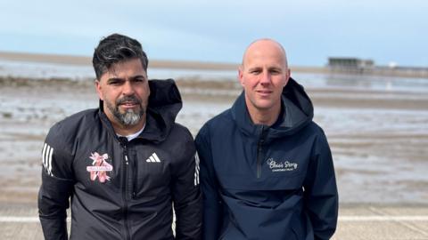Sergio Aguiar (left) with black hair and beard wearing a black track suit jacket stands alongside David Stancombe who is bald wearing a navy tracksuit jacket. They stand at Southport seafront with the pier in the distance.