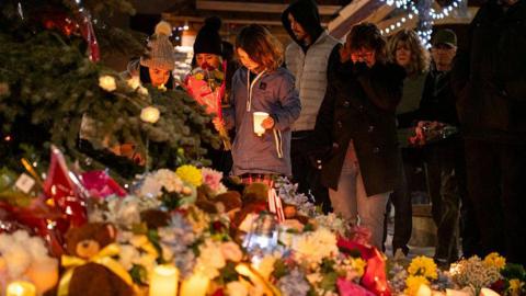 Community members mourn during a candlelight vigil for the victims of Tumbler Ridge Secondary School where a mass shooting took place a day earlier in the small town of Tumbler Ridge, British Columbia, on February 11, 2026. A pile of flowers and candles can be seen at the forefront of the image, while several people look down in the background towards the vigil with sombre expressions. 