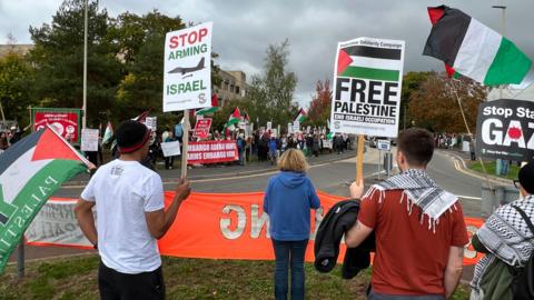 A large group of protesters holding Palestine flags, banners, and signs with slogans such as "Free Palestine" and "Stop Arming Israel" stand either side of a quiet road in a business park-type environment with trees and lamp posts. There is a cloudy, grey sky above.