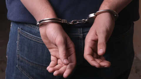 A close-up of the back of a person's hands in handcuffs. The person is wearing blue jeans and a blue T-shirt.