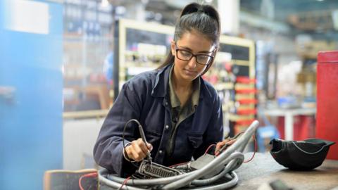 A young woman sits in a factory and uses a pen-like tool to work on a piece of electrical equipment. She has long black hair tied in a ponytail and glasses, and she is wearing a dark blue overall.