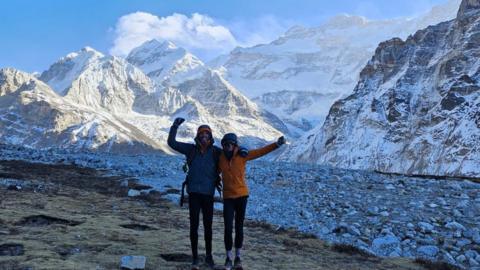 Rik and Liv celebrate with the mountains behind them. They have their arms around each other's shoulders and are lifting their other arms into the air. Rik is wearing a dark blue coat and Olivia has a orange coat. They both have gloves and dark trousers on with their coat hoods over their heads.