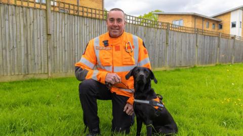A man in an orange hi-vis jacket is kneeling next to a black labrador at a garden.