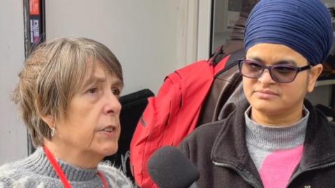 Woman on left wearing a grey jumper, has brown hair and a red lanyard talking into a microphone. Woman on right wearing a hijab, glasses, grey jumper and black cardigan listens in.