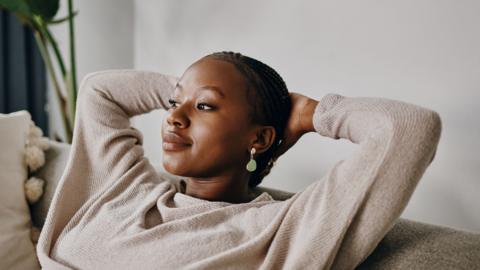 A young woman relaxes on her sofa with both of her hands behind her head.