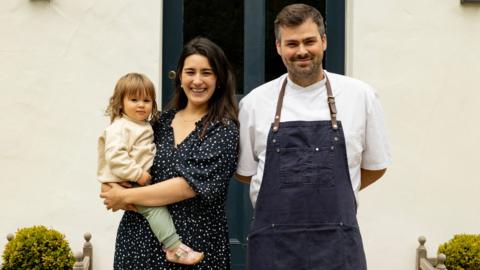Matilda and Tom Tsappis standing outside Killiecrankie House. Matilda has shoulder length dark brown hair and is wearing a black dress with white spots and is holding a toddler on her hip. Tom has short brown hair and a trimmed beard. He is wearing a white chef's jacket and a navy blue apron with leather neck strap.