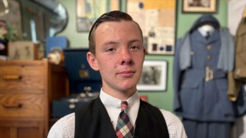 A teenage boy in 1940s dress sitting in front of memorabilia from that era.